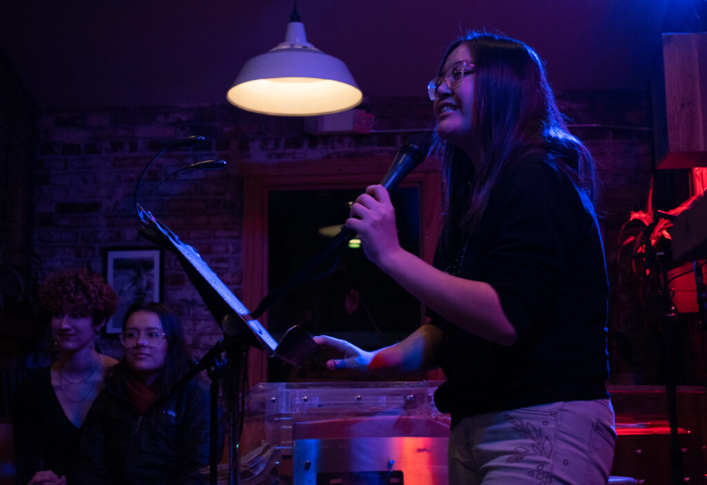 A speaker in glasses and a sweatshirt holds the mic in front of a music stand in a dimly-lit venue. 