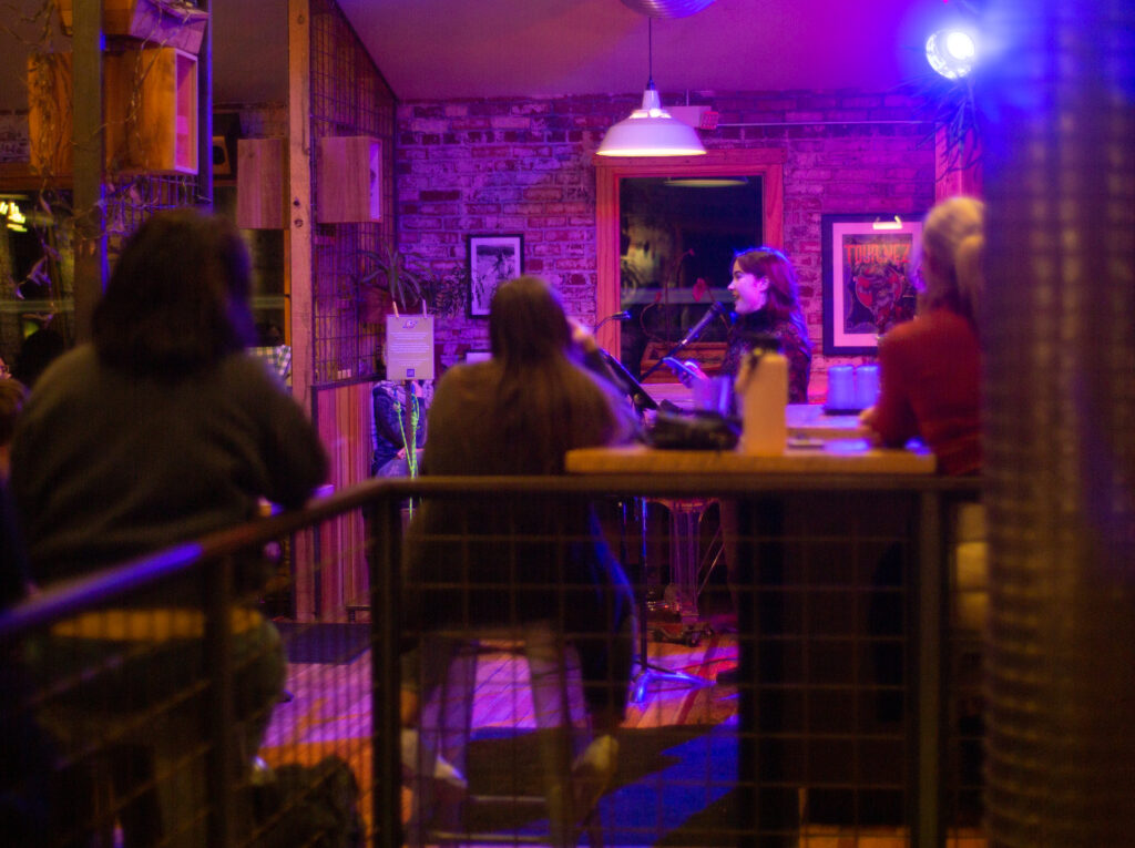 A speaker stands at a microphone on the 2nd floor of the Laughing Planet. Three viewers are visible, with their backs turned to the camera.