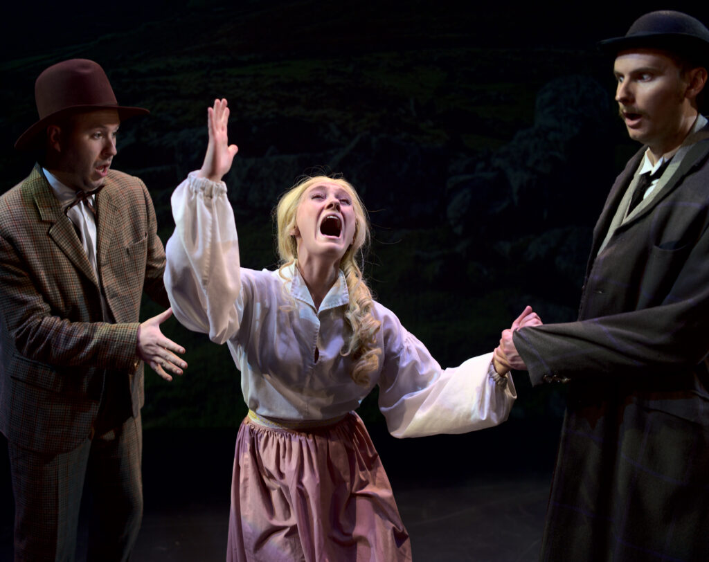 Three actors on stage. One in the center, wearing a blonde wig, shouts through a frantic gesture.