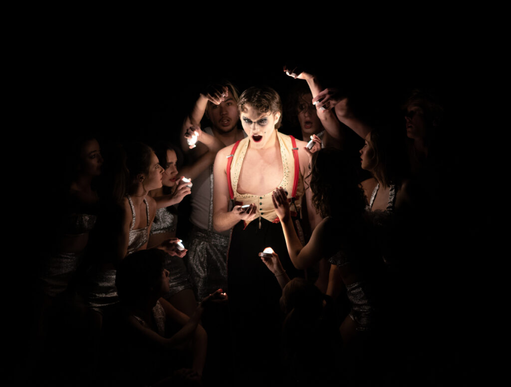 An actor mid-musical number, surrounded by ensemble members with handheld lights. 