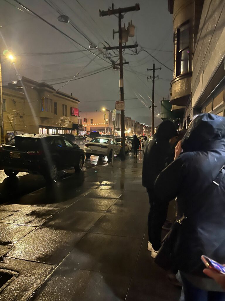 A view on a rainy street. The sidewalk reflects the street lights, and concertgoers in coats stand in the foreground. 