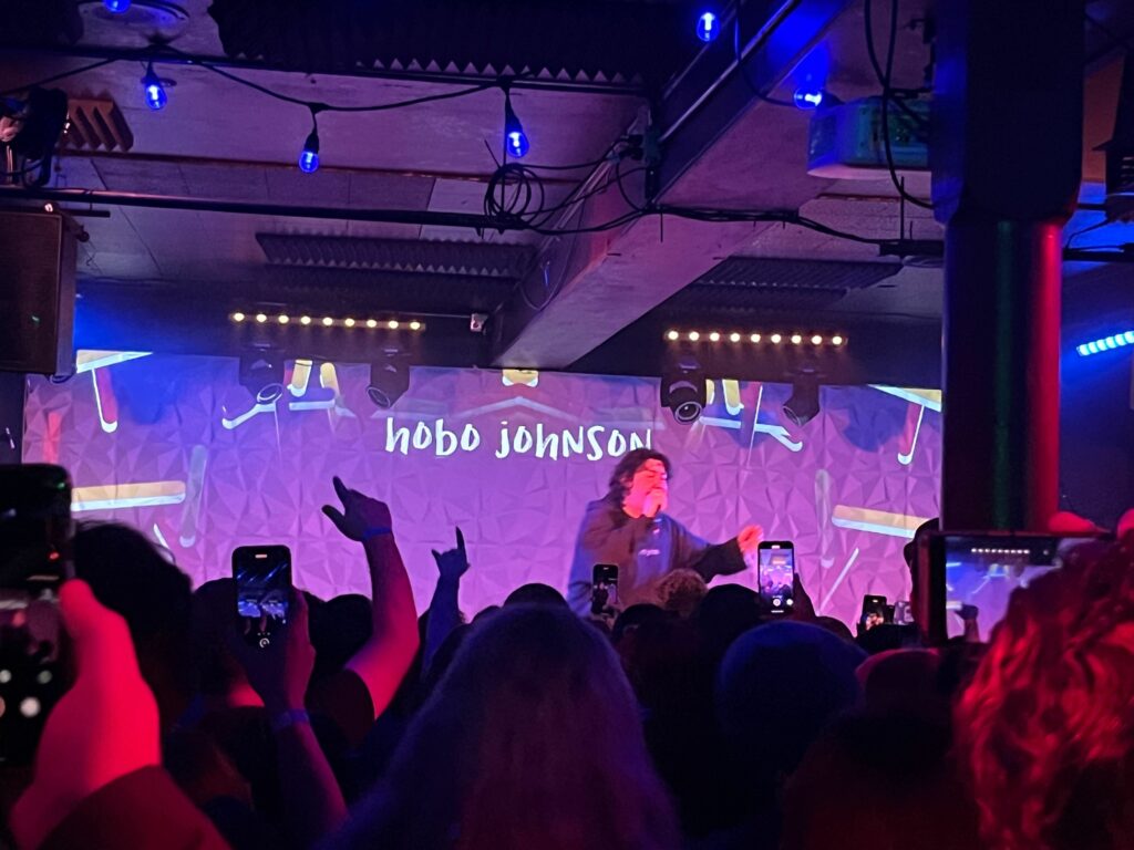 A packed concert venue. An artist at the front is lit from behind with an LCD display that reads "Hobo Johnson".