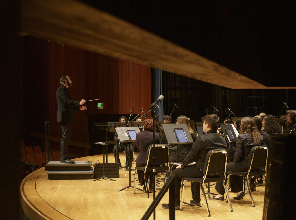 View of the stage from stage left, with the conductor to the left and musicians arranged in rows to the right. 