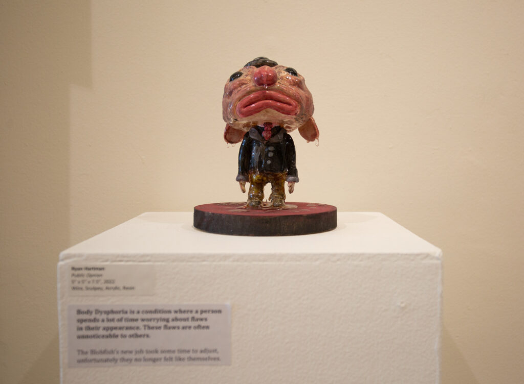 A small, clay sculpture of a blobfish in a suit. It stands atop a small white podium, a description pasted to the left. 