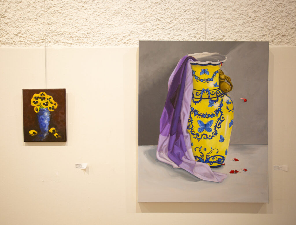 Two oil paintings on a gallery wall. One is of a vase holding yellow flowers, the other of an intricate vase with only a bolt of fabric still in it. 