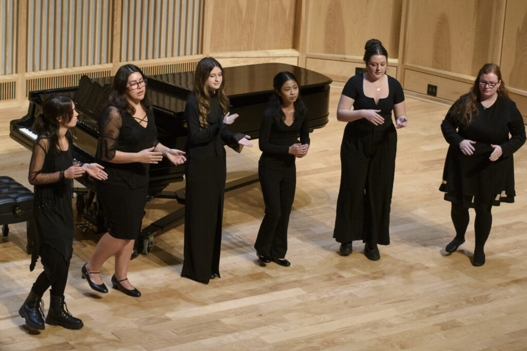 A group of six singers in black dresses stand on a recital hall stage.
