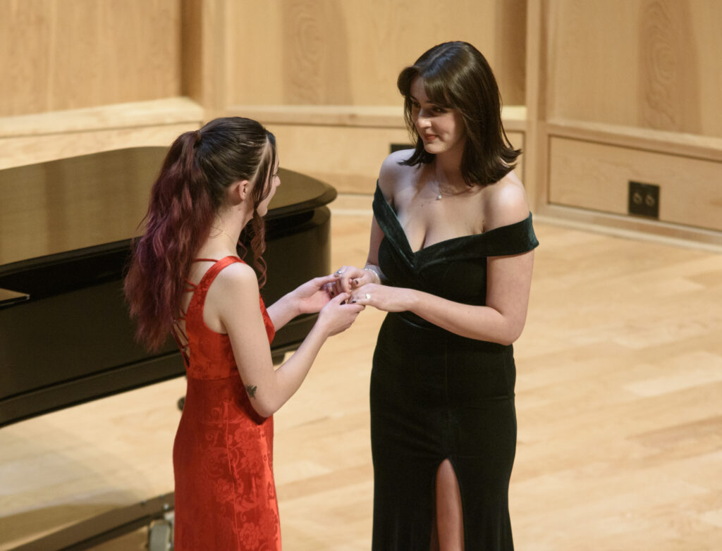 Two singers in a duet with hands clasped. The performer on the white is in a red dress, the performer on the left is in green.