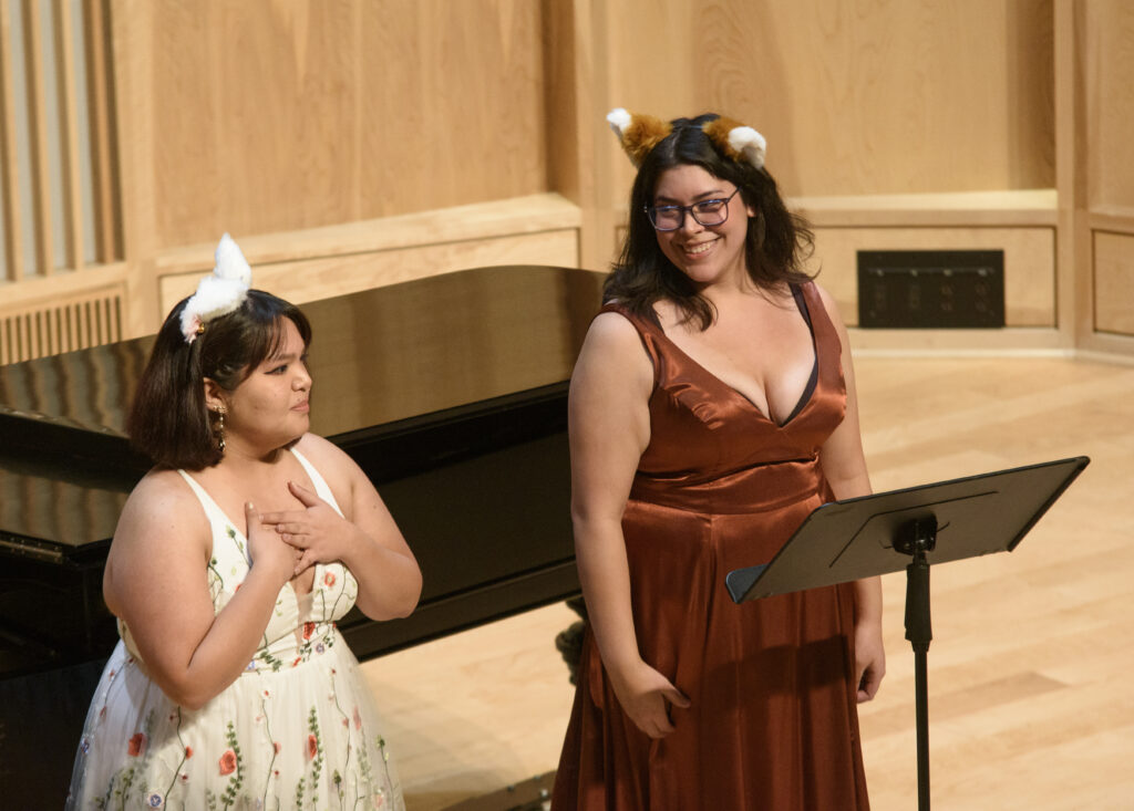 Two singers in dresses and cat ears perform in front of a piano.