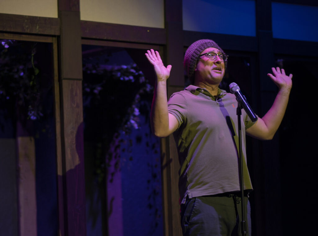 A guy in a polo shirt and a beanie stands at a microphone, looking distressed or excited or both. 