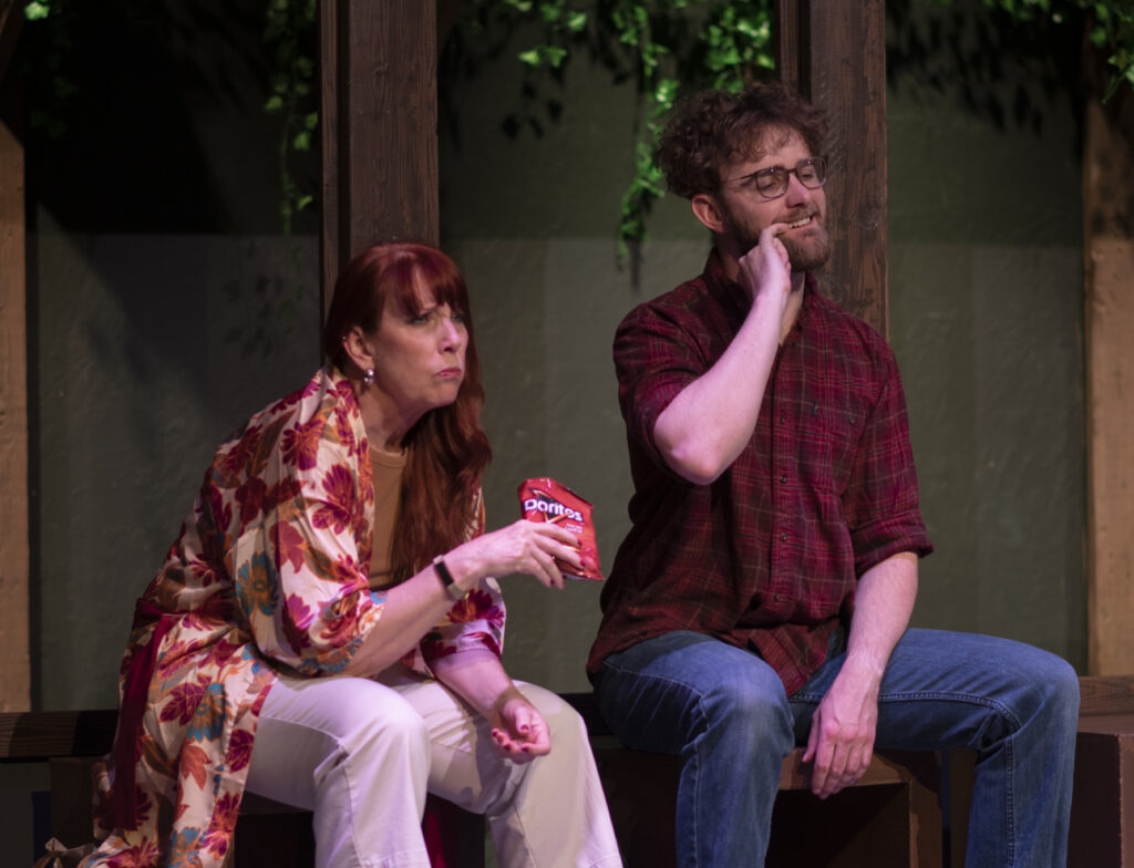 Two people sit hunched on stage. One is mid-snack and the other picks idly at his teeth. 