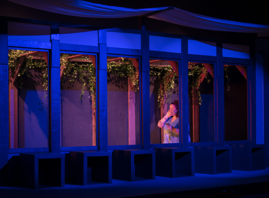 Lit by blue light, an actress holding a phone claps a shocked hand over her mouth. 