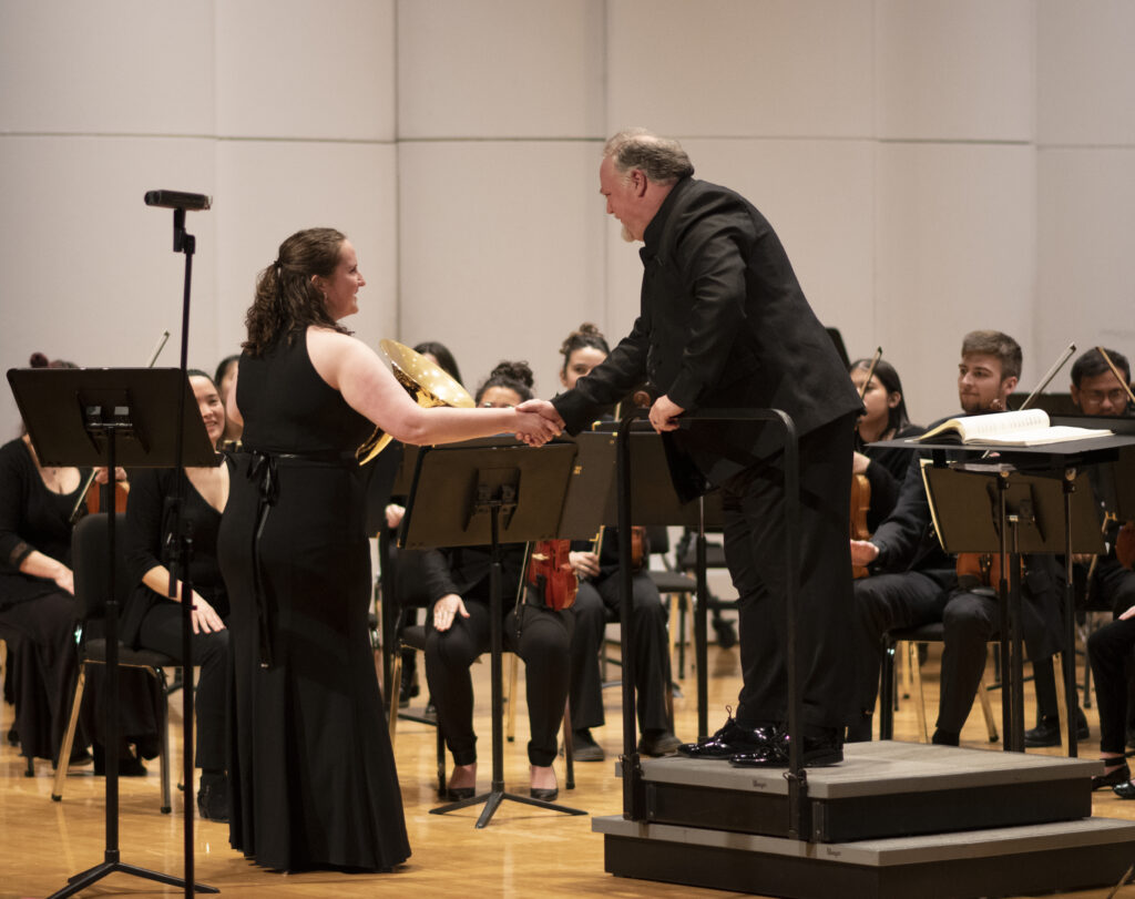 Against a seated orchestra, a conductor shakes the hand of a player with a horn.