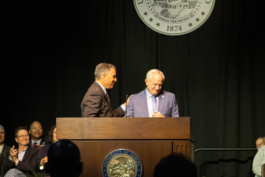 Brian Sandoval and Jeff Thompson greet each other at the podium at the state of the university while the deans sit behind him on the stage.