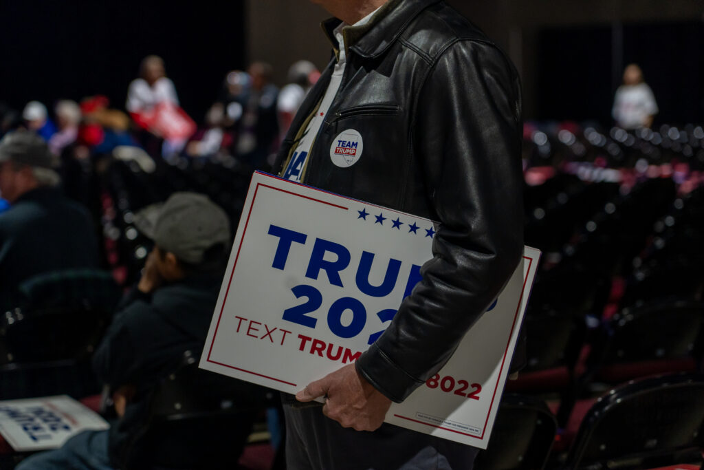 Man in leather jacket holds "Trump 2024" sign at rally.