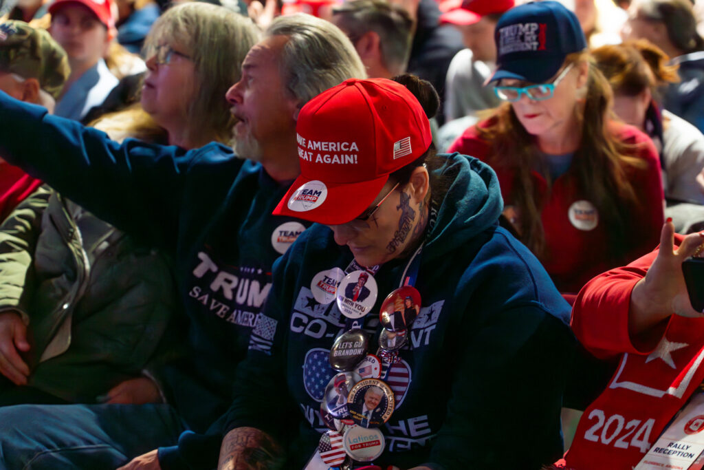 Women looking down texting wearing lots of Trump merchandise and Trump pins, including a "Make America Great Again" red hat.