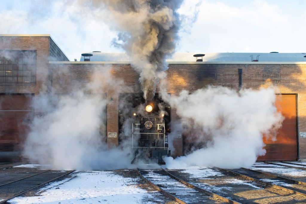 A black steam locomotive is exiting a building. There is a lot of smoke and steam escaping from the engine. 