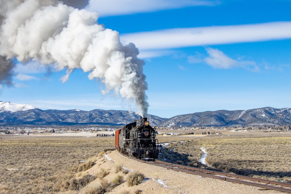 A black steam locomotive is releasing big, puffy clouds of steam. There are snowy mountains in the background. 