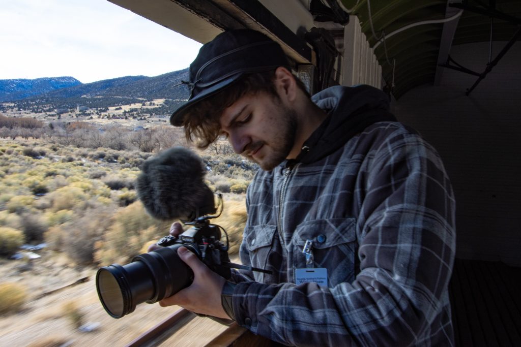 A young man with a camera is leaning out the window of a train.