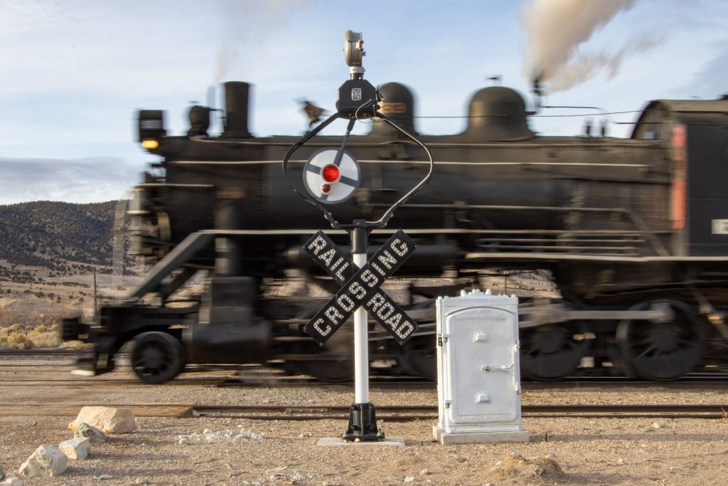 There is a railroad crossing sign with a black steam locomotive passing behind the sign. 