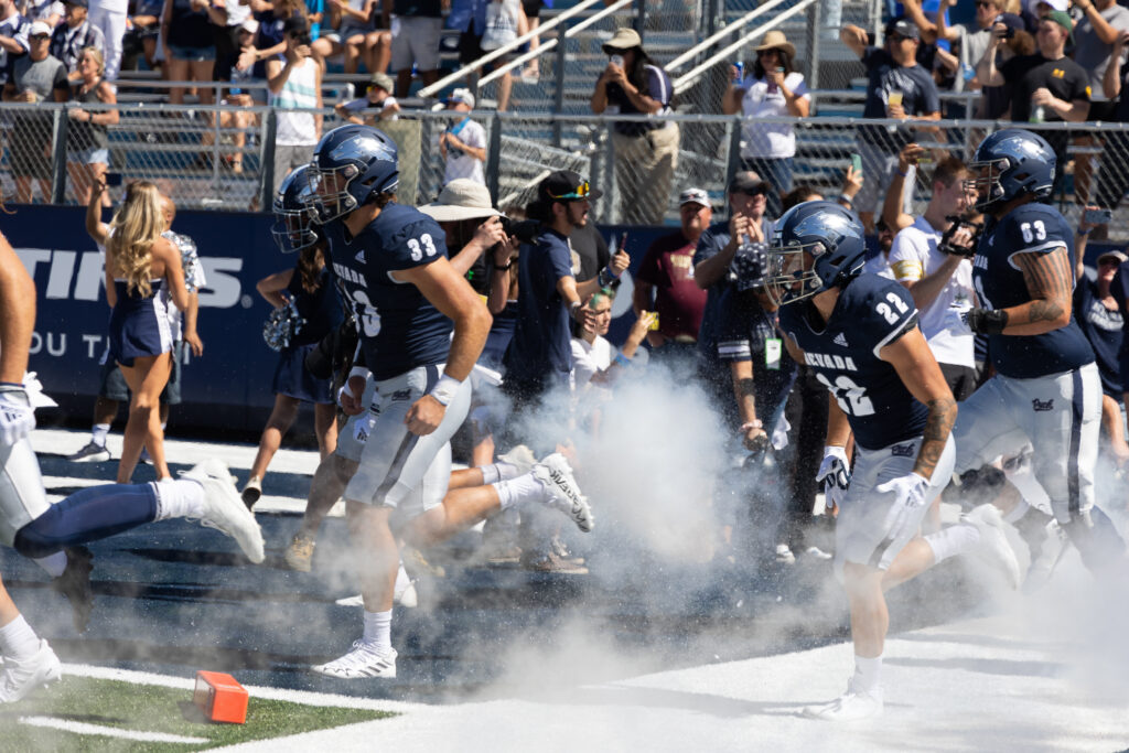 Nevada football team members run out onto the football field with steam surrounding them