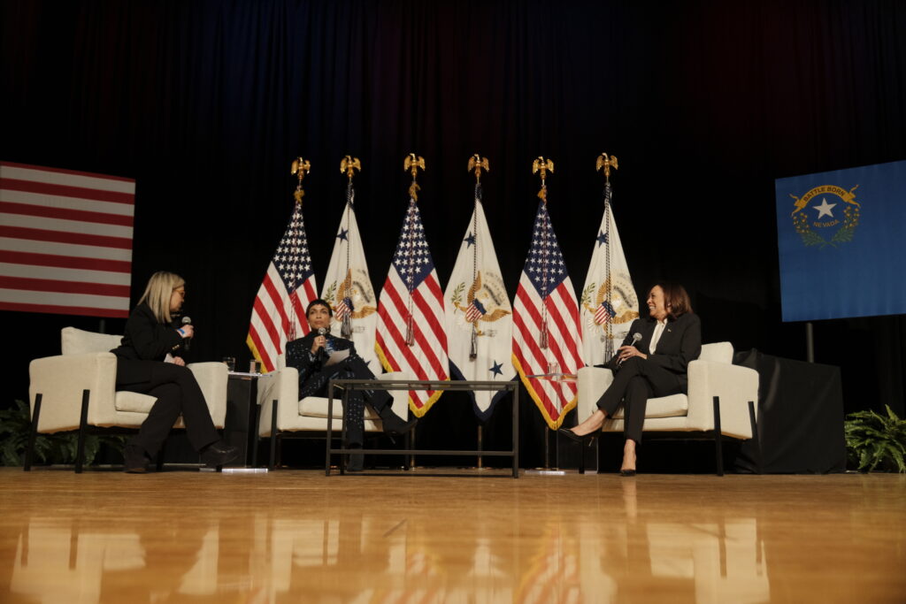 Hillary Schieve, Rosario Dawson and Kamala Harris speak on a stage sitting in white chairs.