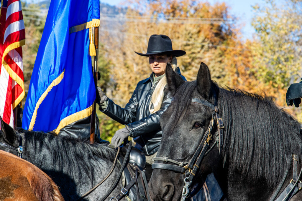 Woman in a black leather jacket and black hat sits on top of a black horse, holding a Nevada flag.