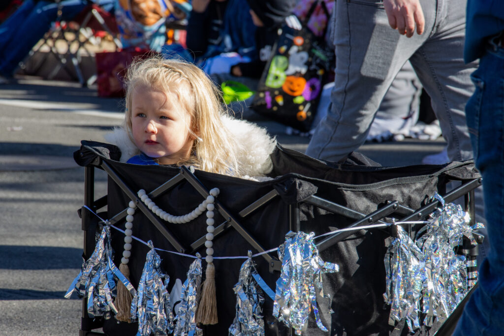 Little girl sits in a wagon with pom poms looking out at the parade attendees.