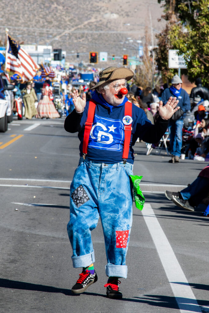 Man wearing a brown hat, clown nose, and red suspenders waves with both hands walking down the street.
