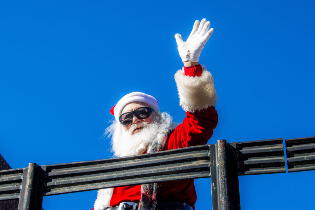 Santa wears sunglasses and his classic red coat and hat, waving.