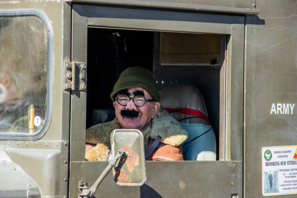 Man wearing Groucho glasses and green beanie looks out of green army truck window.