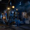 Two locomotives are positoned parallel to each other inside a dark building. One has steam coming out of it, and a man is posing for photos on the front.