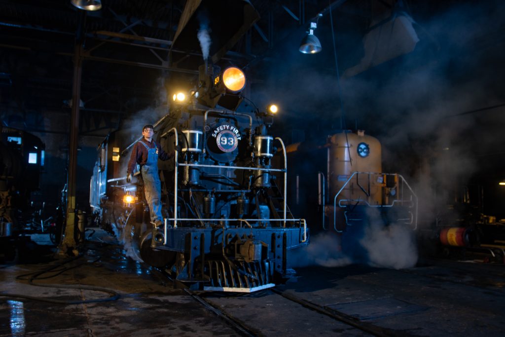 Two locomotives are positoned parallel to each other inside a dark building. One has steam coming out of it, and a man is posing for photos on the front.