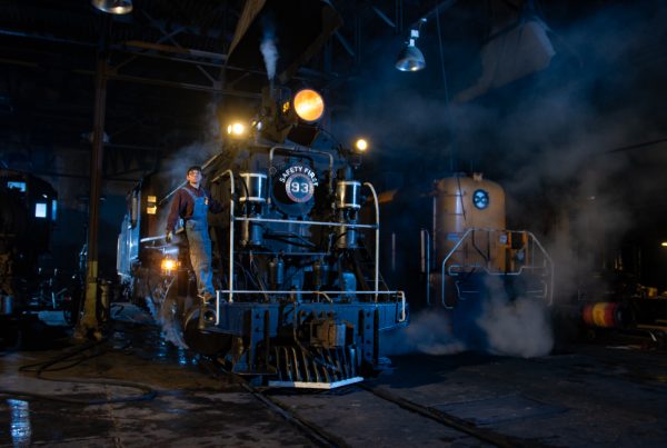 Two locomotives are positoned parallel to each other inside a dark building. One has steam coming out of it, and a man is posing for photos on the front.