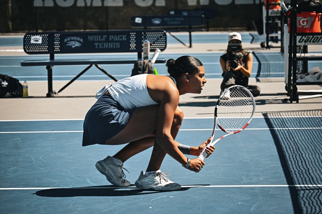 Senior women's tennis player Kennedy Robinson squats down with her eye on the ball during a match.