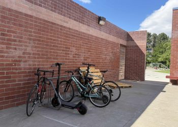 A full bike rack with bikes and scooters. A brick wall is behind the bike rack.