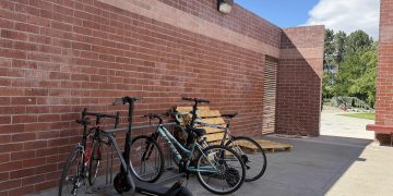 A full bike rack with bikes and scooters. A brick wall is behind the bike rack.
