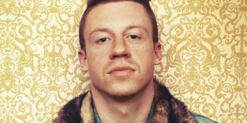 Macklemore stands against a wallpaper background.