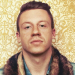 Macklemore stands against a wallpaper background.