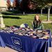 A student sat behind a table with a cloth that reads, "ASUN." She is holding up a wolfpack sign and has candy bars laid out in front of her on the table. She is outside.